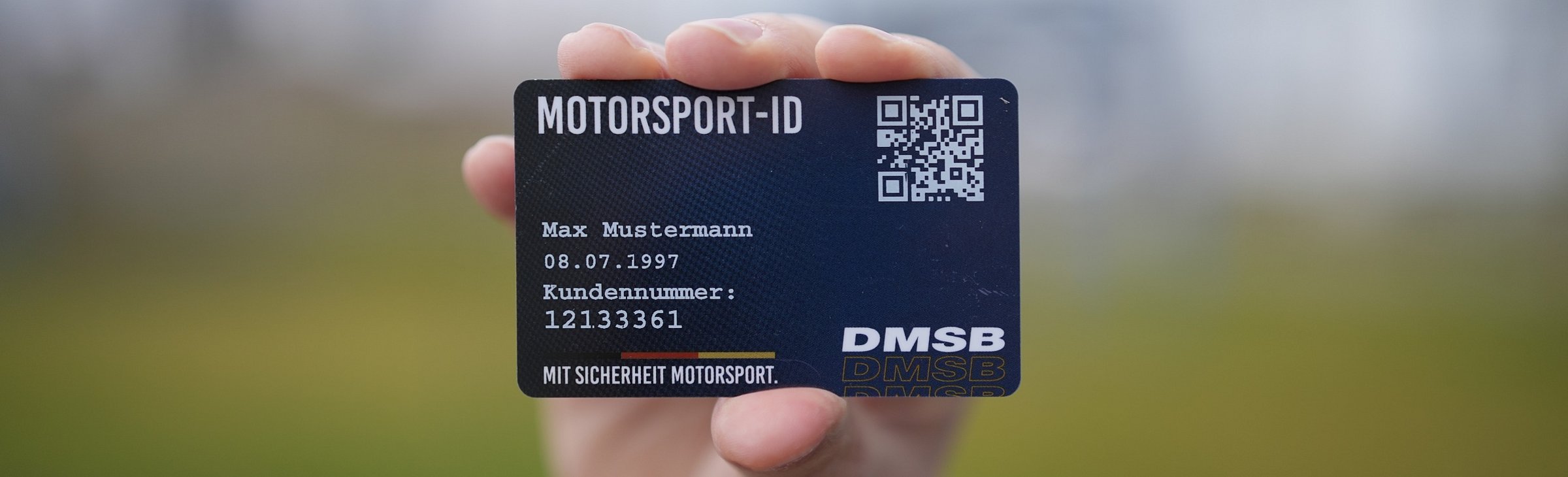 DMSB Motorsport-ID Max Mustermann Eine dunkelblaue Motorsport-ID im Scheckkartenformat mit QR-Code, Daten zu Max Mustermann und dem DMSB Logo. Hintergrund blurred draußen vor einer grünen Wiese und grauem Himmel.