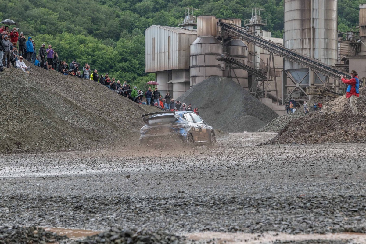 Hunsrück Rallye 2024 Ein Rallyefahrzeug fährt in einer Schotterkurve auf der Rennstrecke bei der Hunsrück Rallye 2024. Am Rand Zuschauer und ein Fotograf.