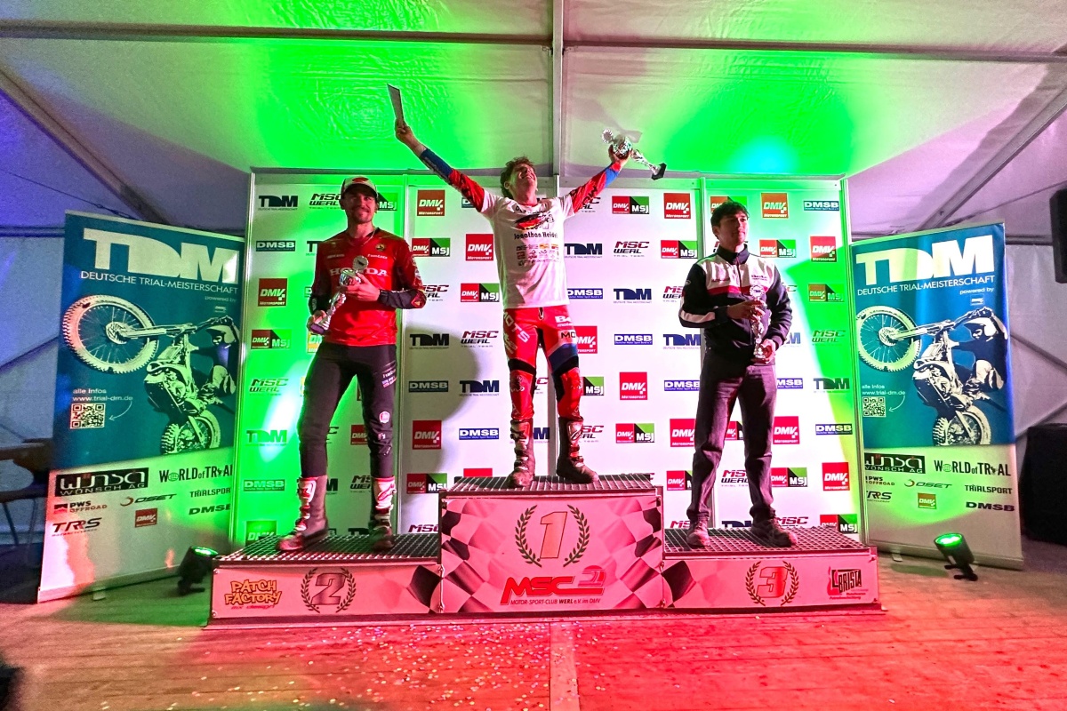Jonathan Heidel feiert auf dem obersten Podestplatz seinen Sieg beim Finale der Deutschen Trial-Meisterschaft 2026 in Werl. Jonathan Heidel feiert auf dem obersten Podestplatz seinen Sieg beim Finale der Deutschen Trial-Meisterschaft 2026 in Werl. Neben ihm stehen Florian Stephan auf Rang zwei und Rodney Bereiter auf Rang drei mit ihren Pokalen vor der Sponsorentafel.