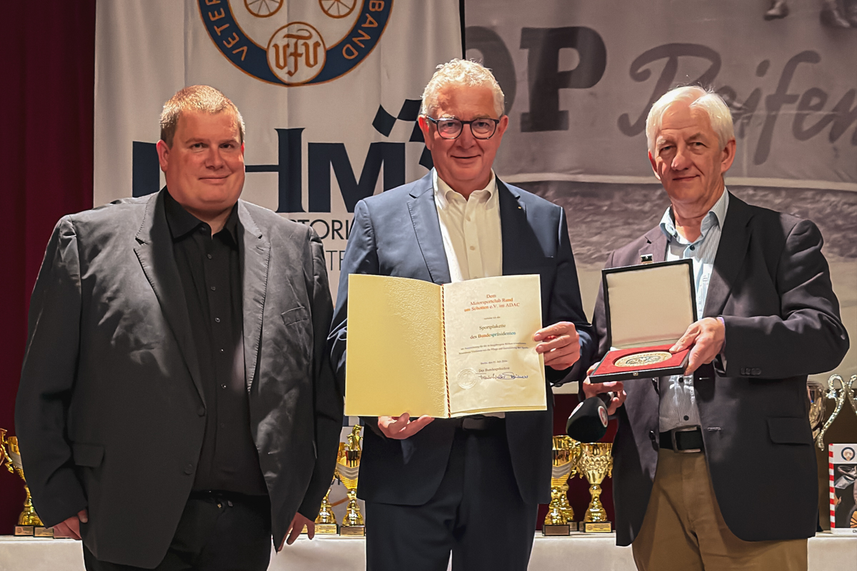 Drei Männer stehen nebeneinander; der Mann in der Mitte hält eine Urkunde in der Hand, und der Mann rechts hält eine geöffnete Prämienbox, in der sich eine Medaille befindet.