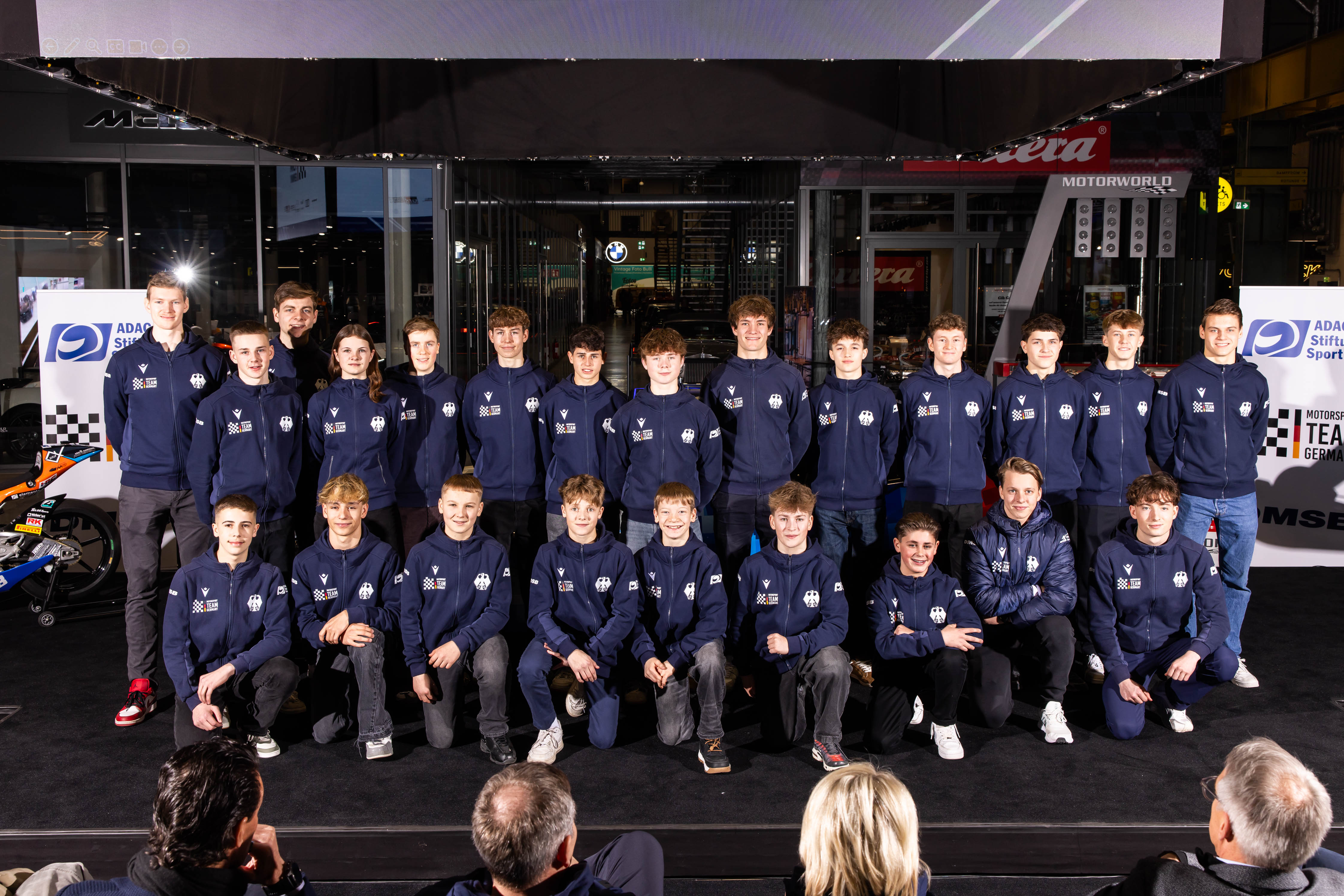 Eine Gruppe von 22 jungen Leuten in passenden blauen Jacken stellt sich auf einer Bühne für ein Gruppenfoto zusammen.